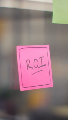 Vertical-Video-Close-Up-Of-Woman-Putting-Sticky-Note-With-ROI-Written-On-It-Onto-Transparent-Screen-In-Office